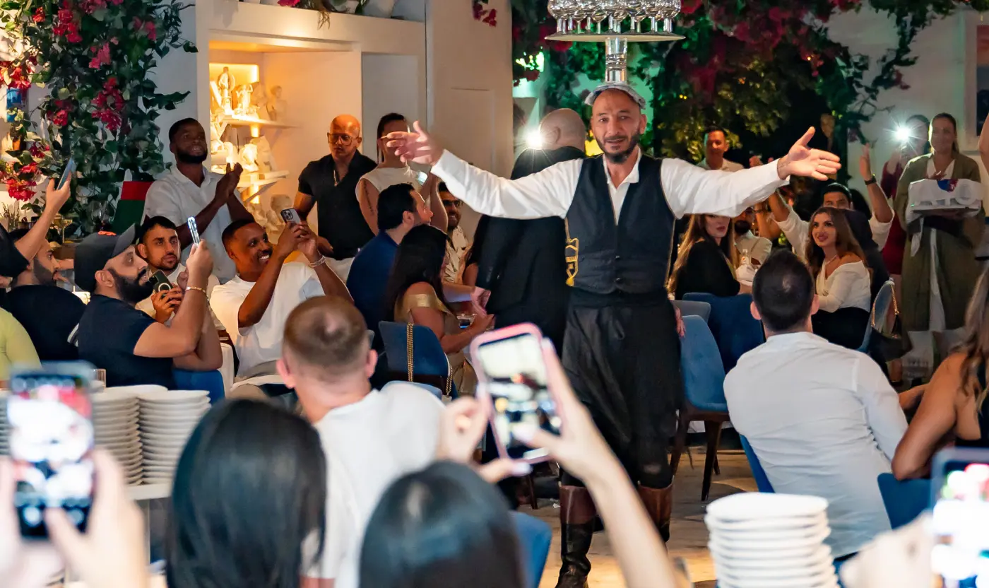 A traditional Greek restaurant performer balancing a tower of glasses on his head amidst dining guests.
